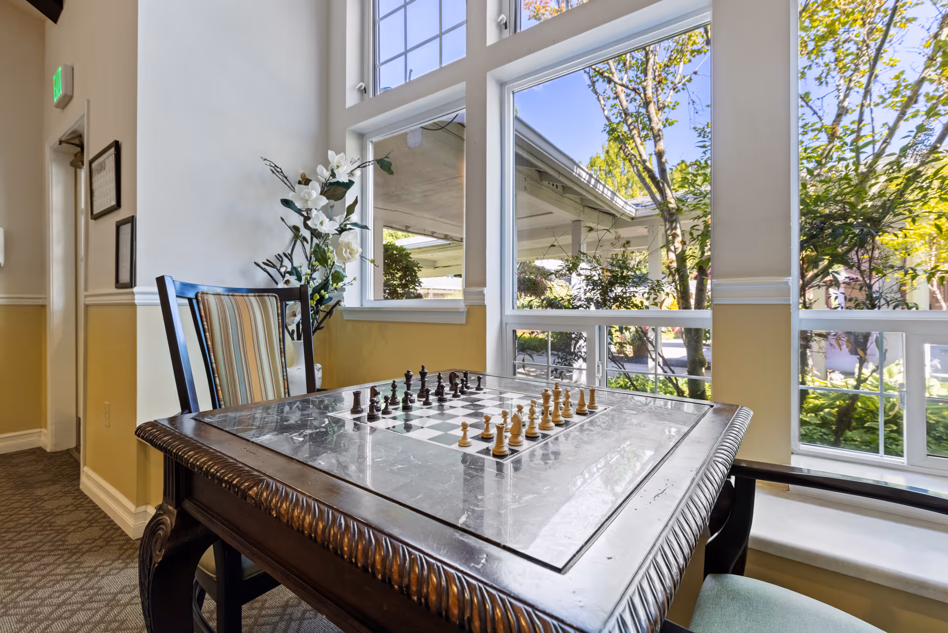 A chess table with a chessboard and pieces set up in a well-lit room with large windows showing greenery outside. There are two chairs around the table, one with striped upholstery and the other with a solid cushion. A decorative plant with white flowers is placed near the wall.