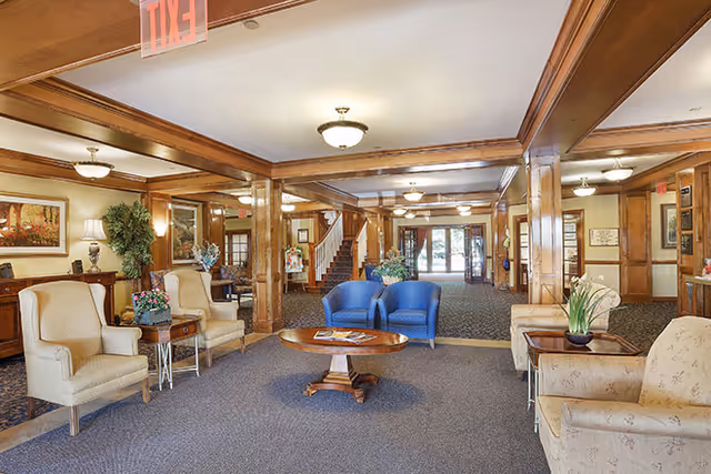 A spacious and well-lit common area in a senior living facility with comfortable seating including beige armchairs, blue chairs, and a beige sofa. The room features wooden columns and trim, a central wooden coffee table with magazines, decorative plants, framed artwork on the walls, and ceiling lights. A staircase and glass doors leading outside are visible in the background.