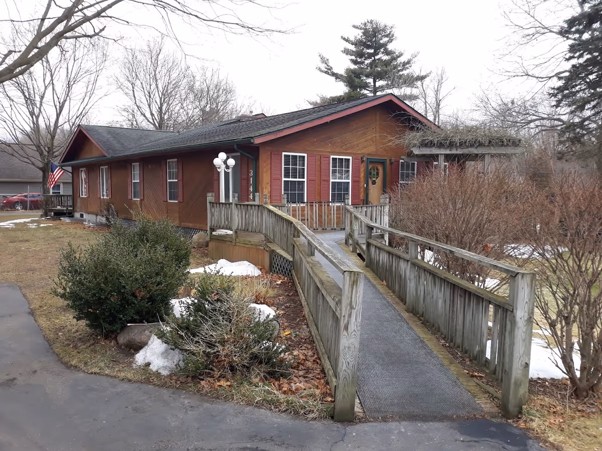 Front exterior of a single-story brown wooden building with an accessibility ramp leading to the entrance, surrounded by leafless trees and patches of snow.
