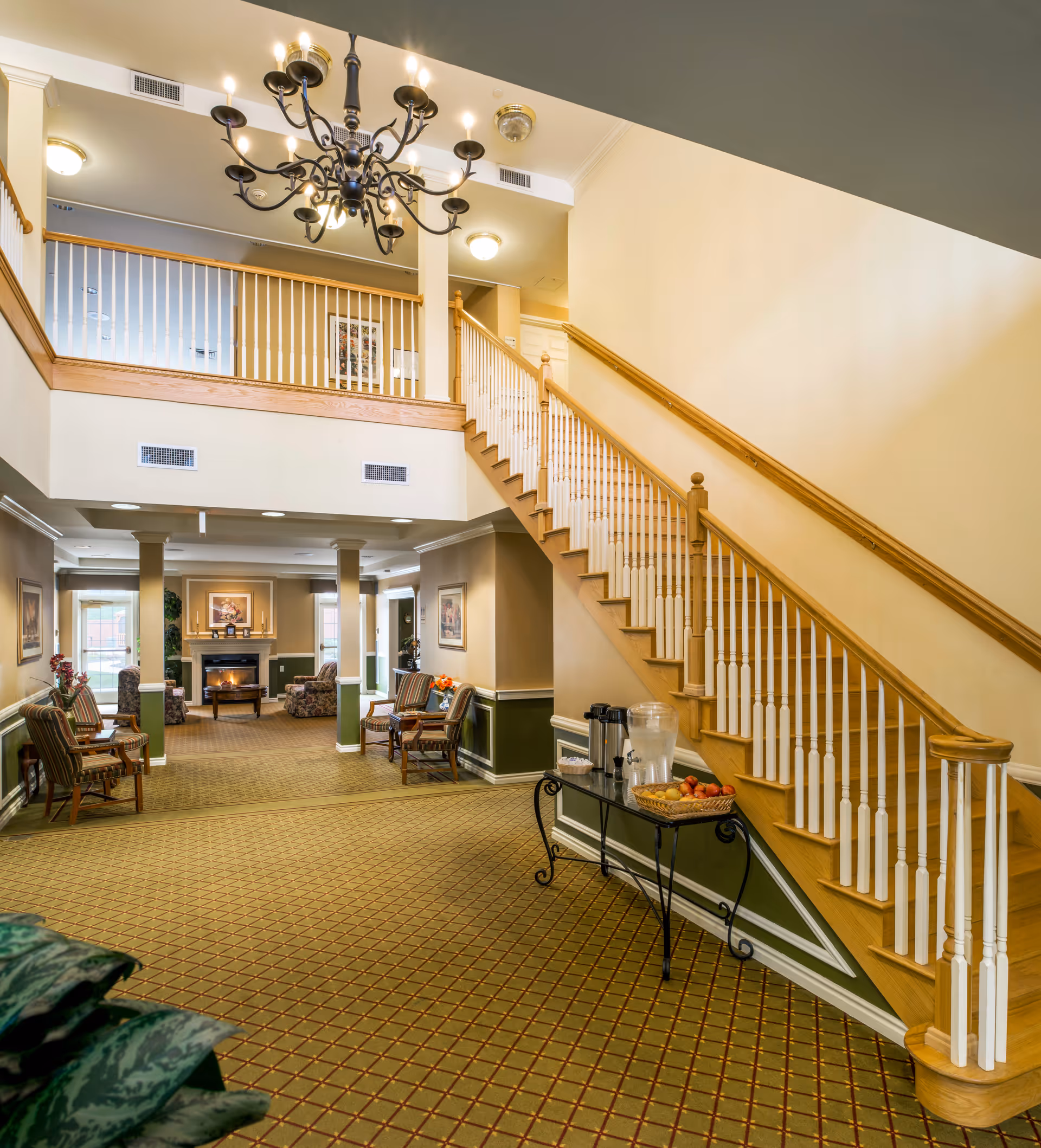 Bright assisted-living lobby with a wooden staircase, chandelier, seating area, fireplace, and a refreshment table.