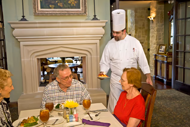 A chef in a white uniform and tall hat serving a dessert to an elderly woman in a red shirt seated at a dining table with two other elderly people. The table is set with glasses of iced tea, plates, and silverware, and there is a fireplace in the background.