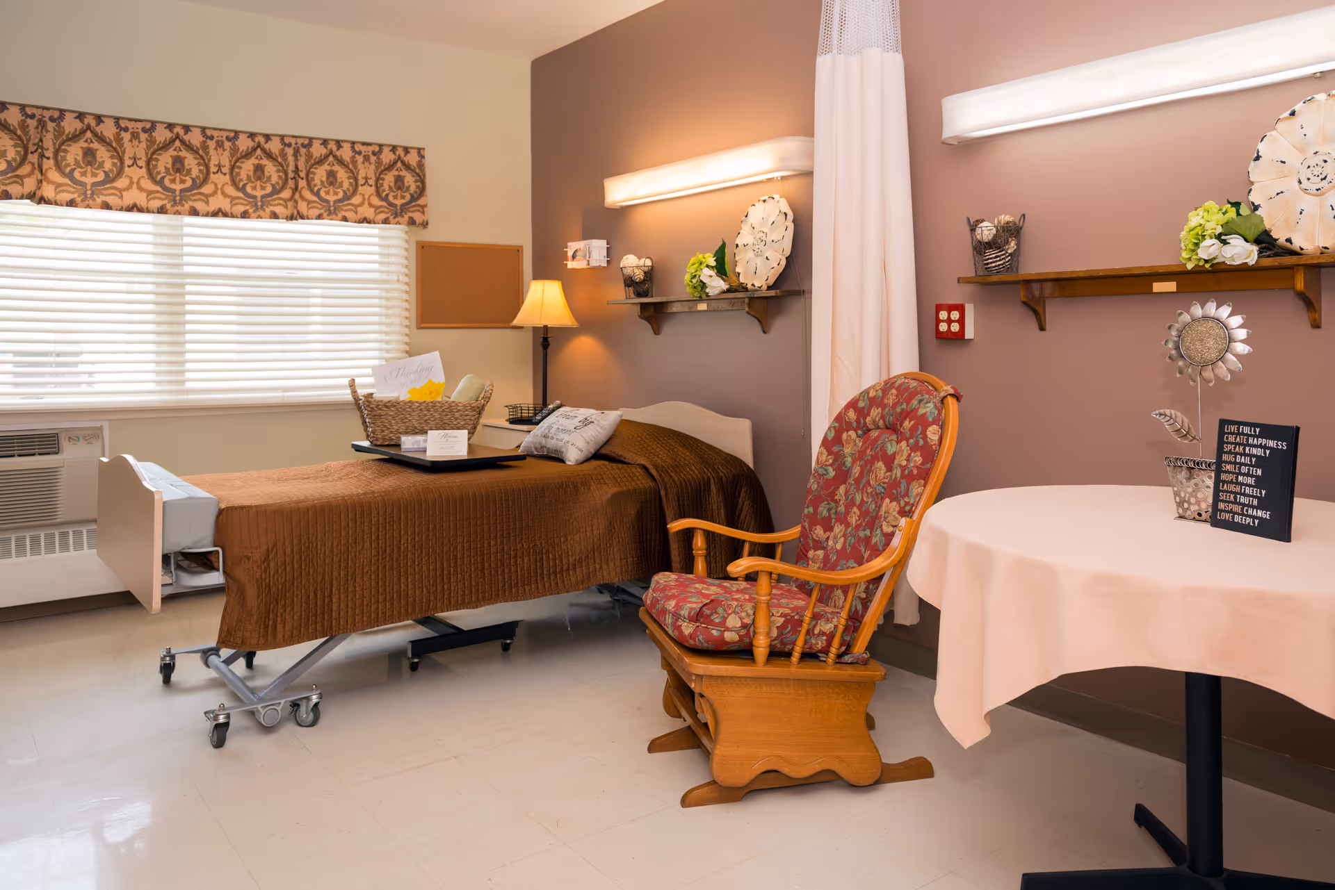 A cozy senior living room with a hospital-style bed covered in a brown blanket, a wooden rocking chair with floral cushions, a round table with a white tablecloth, and decorative items on shelves and the table. A window with blinds and a patterned valance lets in natural light.