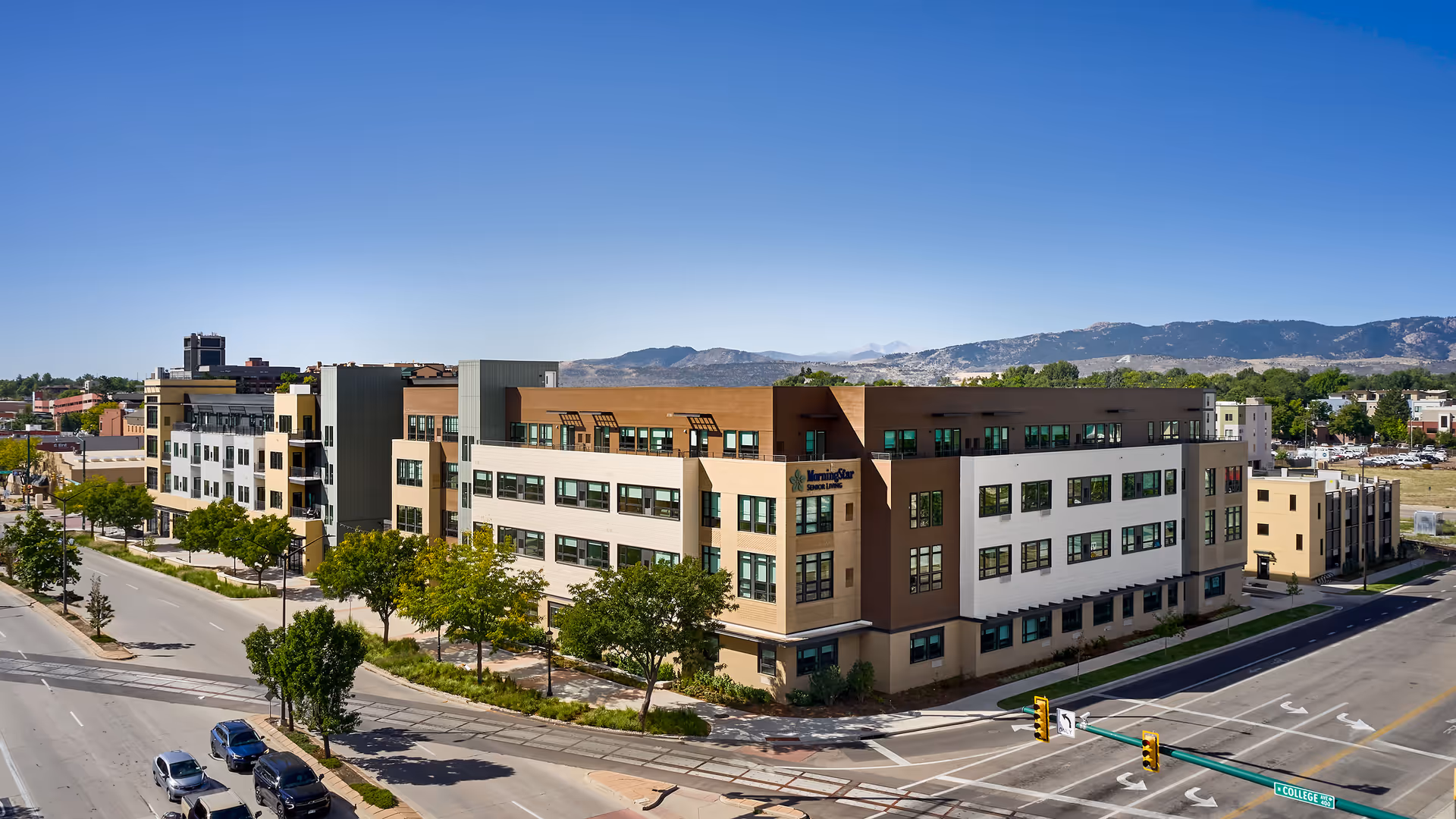 A large, modern four-story senior living facility building with a mix of beige, brown, and white exterior walls, numerous windows, and a sign that reads 'MorningStar Senior Living'. The building is situated at a street intersection with traffic lights and surrounded by trees and other urban buildings. Mountains are visible in the background under a clear blue sky.