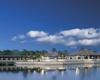 A serene evening view of The Barclay at Sarasota facility with a calm body of water in the foreground reflecting the building and palm trees under a blue sky with scattered clouds.