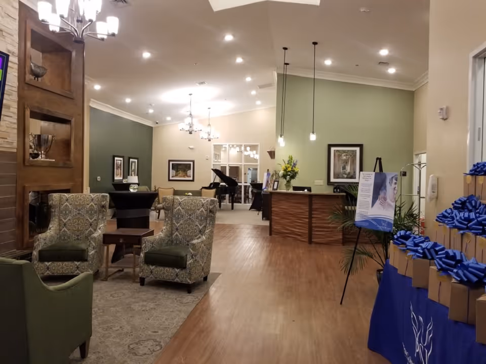 Spacious senior living facility lobby with upholstered chairs, a reception desk, hanging lights, a grand piano, and a table of gift bags.