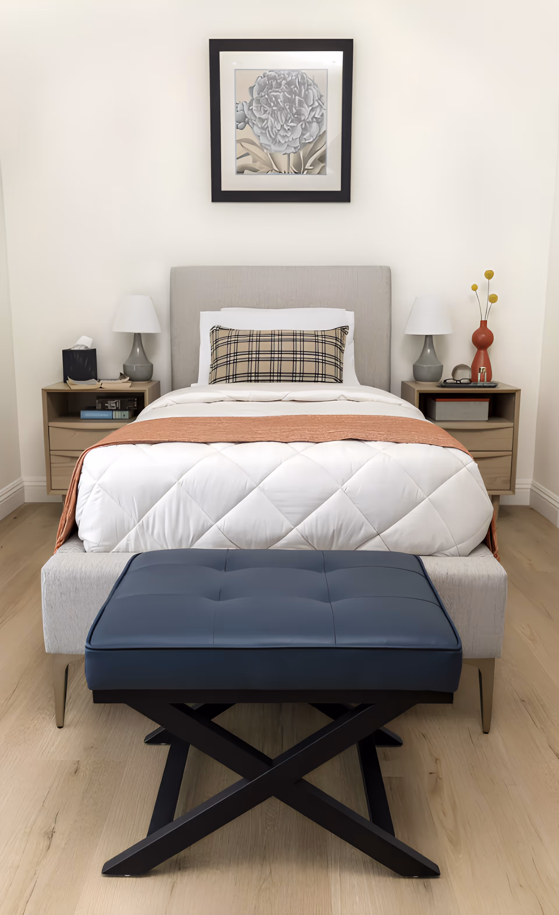 A neatly made single bed with a gray upholstered headboard, white quilted bedding, a plaid decorative pillow, and a rust-colored throw blanket. On either side of the bed are matching nightstands with gray lamps, books, and decorative items. A framed floral artwork hangs on the white wall above the bed. In front of the bed is a dark blue cushioned bench with black crossed legs. The room has light wood flooring.