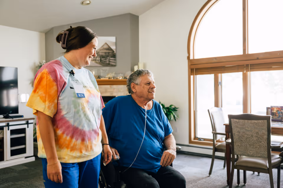 A nurse wearing a colorful tie-dye shirt and an RN badge stands next to an elderly man in a wheelchair who is wearing a blue shirt and using an oxygen tube. They are in a bright room with large arched windows, a dining table with chairs, and a TV in the background.