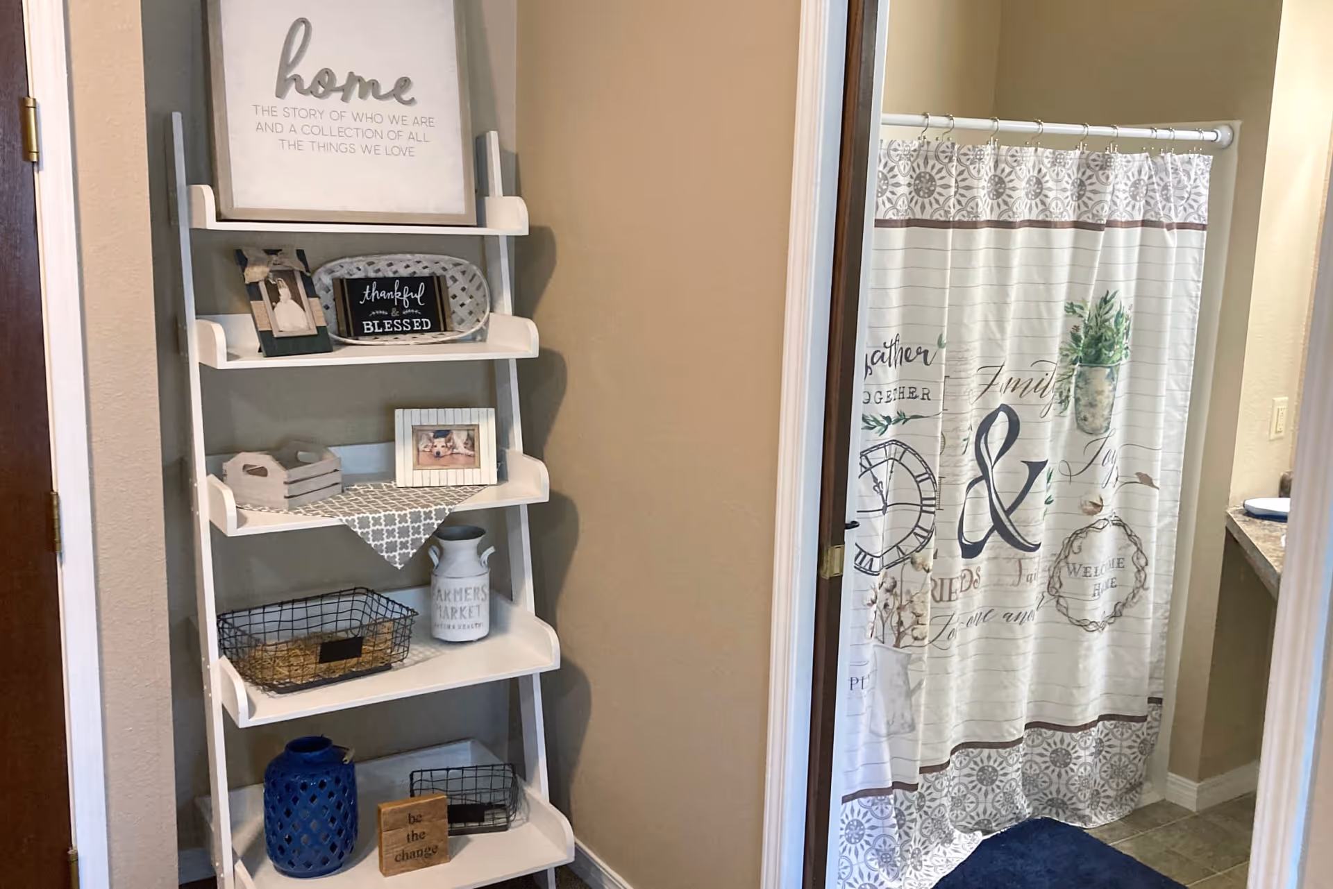 A corner of a room featuring a white leaning shelf decorated with framed photos, decorative signs, baskets, and vases. To the right, there is a bathroom with a shower curtain that has various words and decorative patterns on it, partially visible through an open door.