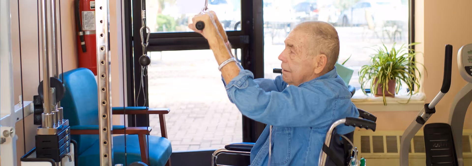 An elderly man in a wheelchair using a pulley exercise machine in a rehabilitation or therapy room with a window, a blue chair, a fire extinguisher, and a potted plant on the windowsill.