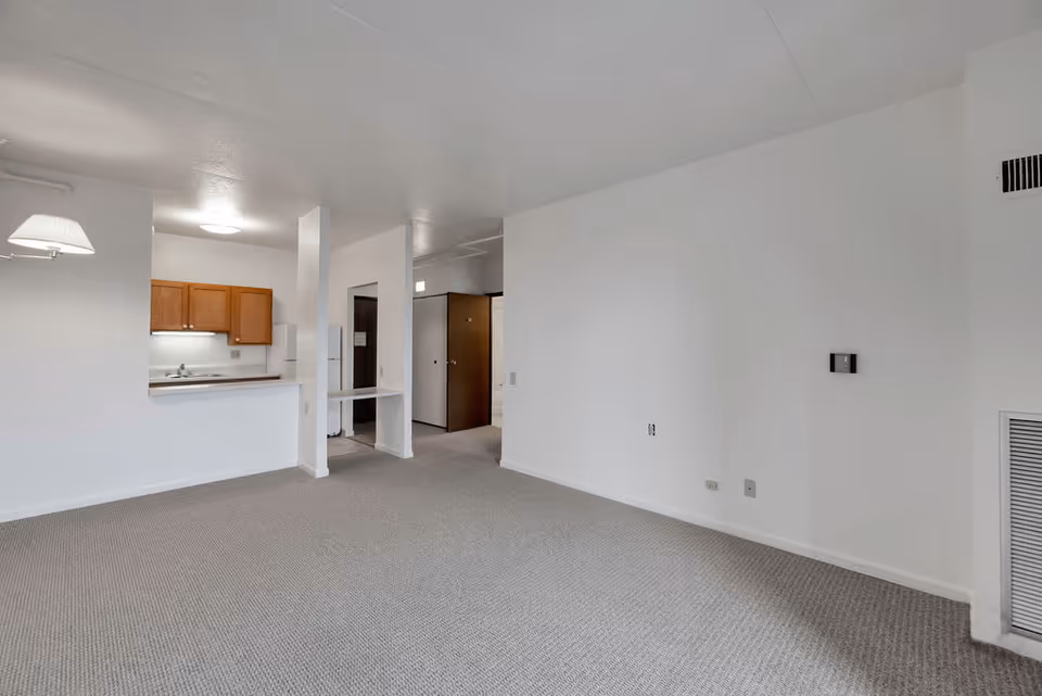 Empty interior room with light gray carpet and white walls. On the left side, there is a small kitchen area with wooden cabinets, a sink, and a refrigerator. The room has a wall-mounted light fixture and an open doorway leading to another room. A thermostat and air vent are visible on the right wall.