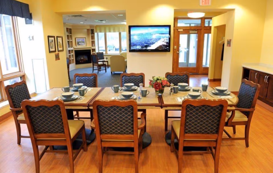 A dining area in a nursing and rehabilitation center with a rectangular table set for eight people. The table has matching chairs with wooden frames and cushioned seats and backs. Each place setting includes a bowl, plate, mug, and utensils. A vase with flowers is in the center of the table. In the background, there is a wall-mounted flat-screen TV displaying a scenic image, a fireplace, and seating area with armchairs. The room has large windows allowing natural light and wooden flooring.