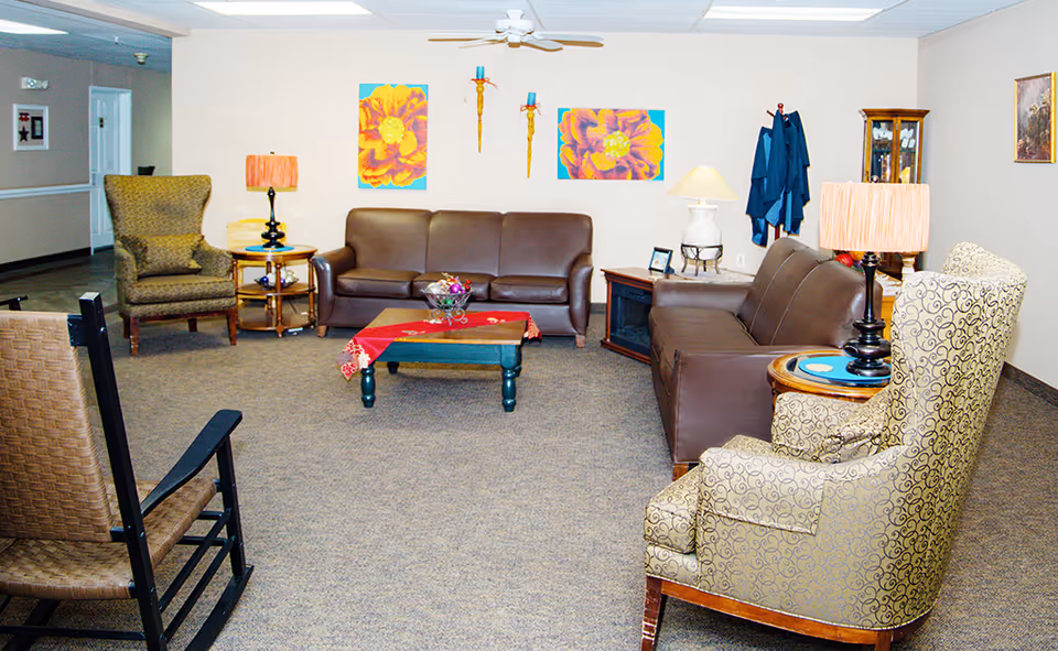A cozy living room area in Peachtree Village Retirement Community featuring two brown leather sofas, two patterned armchairs, a rocking chair, a coffee table with a red runner, two table lamps, colorful flower paintings on the wall, and a coat rack with blue jackets.