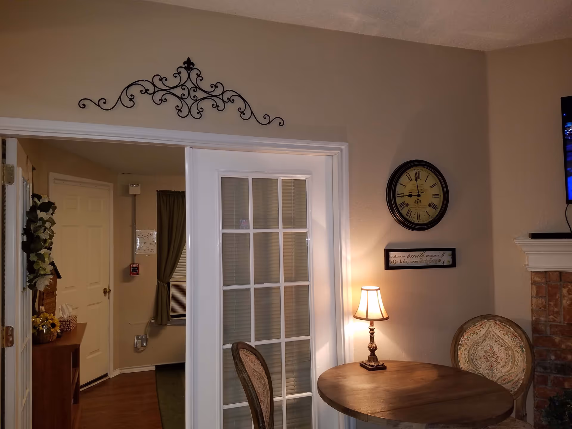 A cozy interior corner of a senior living facility featuring a small round wooden table with two upholstered chairs. A lit table lamp sits on the table. On the wall above the table is a round clock and a framed inspirational quote. To the left, there is a white door with glass panes and a decorative metal wall art above the door frame. A TV is partially visible mounted on the wall to the right, above a brick fireplace.