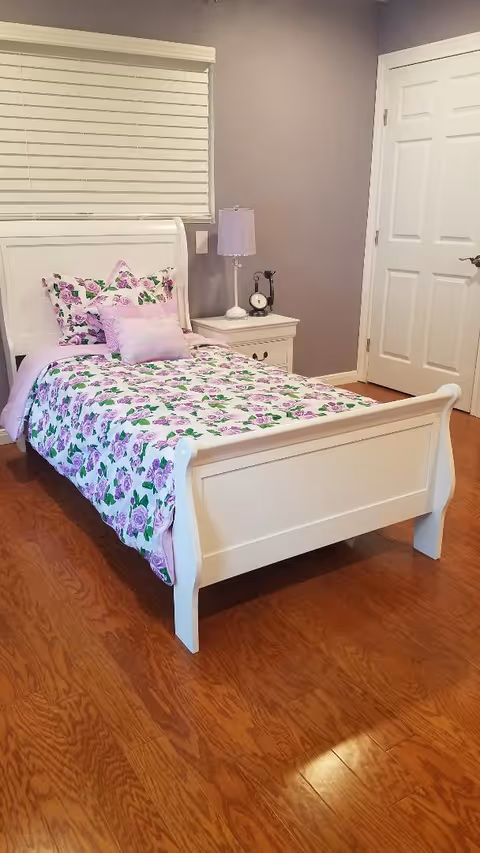 A tidy bedroom with a white single sleigh bed dressed in purple floral bedding and a nightstand with a lamp.