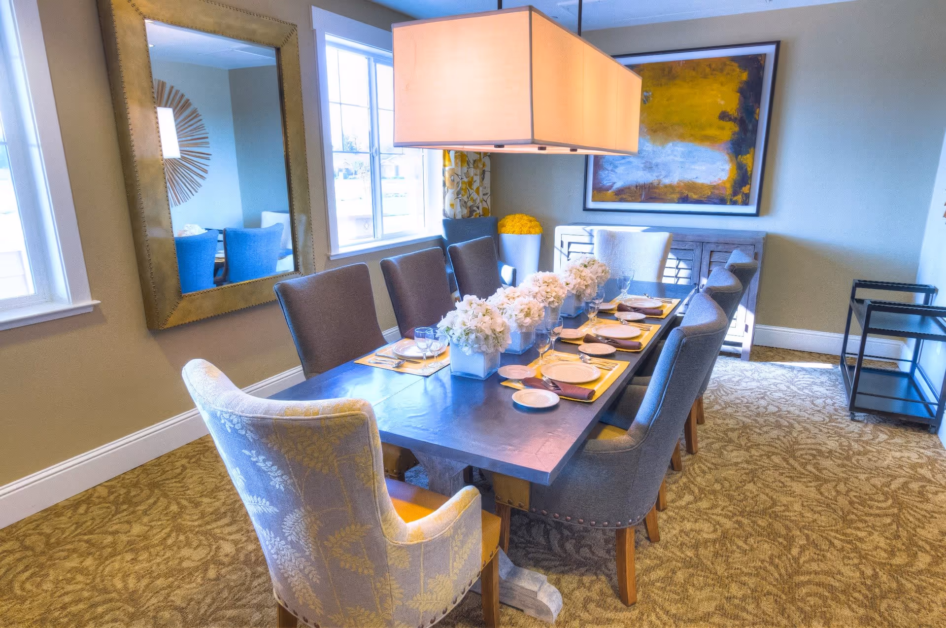 A well-lit dining room with a long rectangular table set for eight people. The table has white floral centerpieces, plates, glasses, and napkins. Surrounding the table are upholstered chairs in shades of gray and beige. A large rectangular pendant light hangs above the table. The room features a large mirror on one wall, a window with patterned curtains, a sideboard with a colorful abstract painting above it, and a carpeted floor with a leafy pattern.