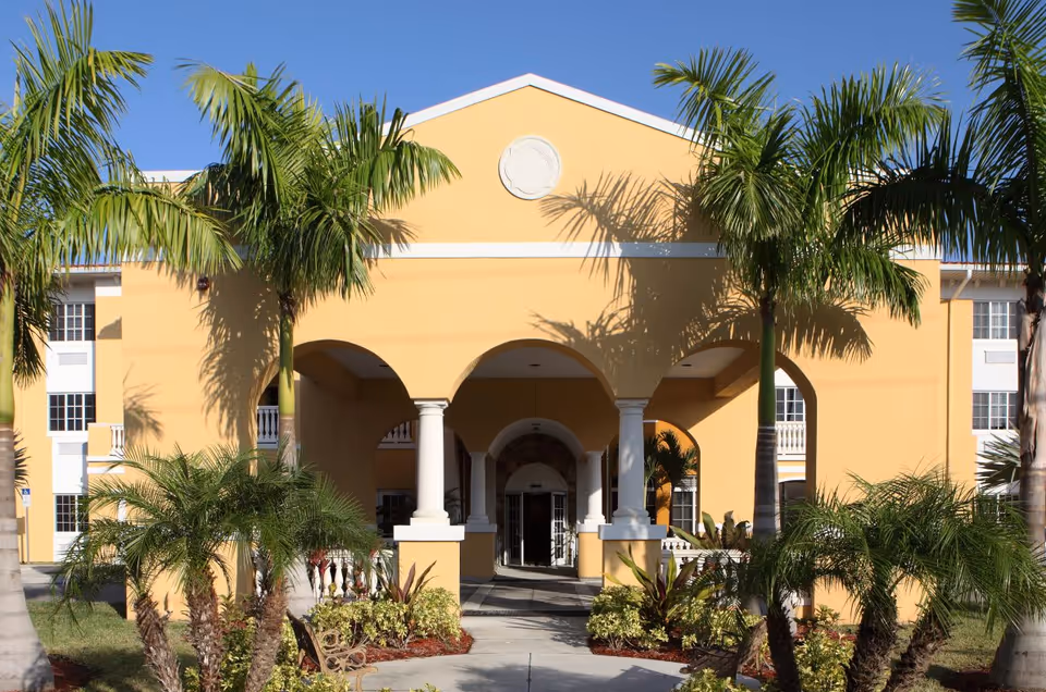 Front entrance of a yellow stucco building with an arched portico, white columns, and palm trees in the landscaping.