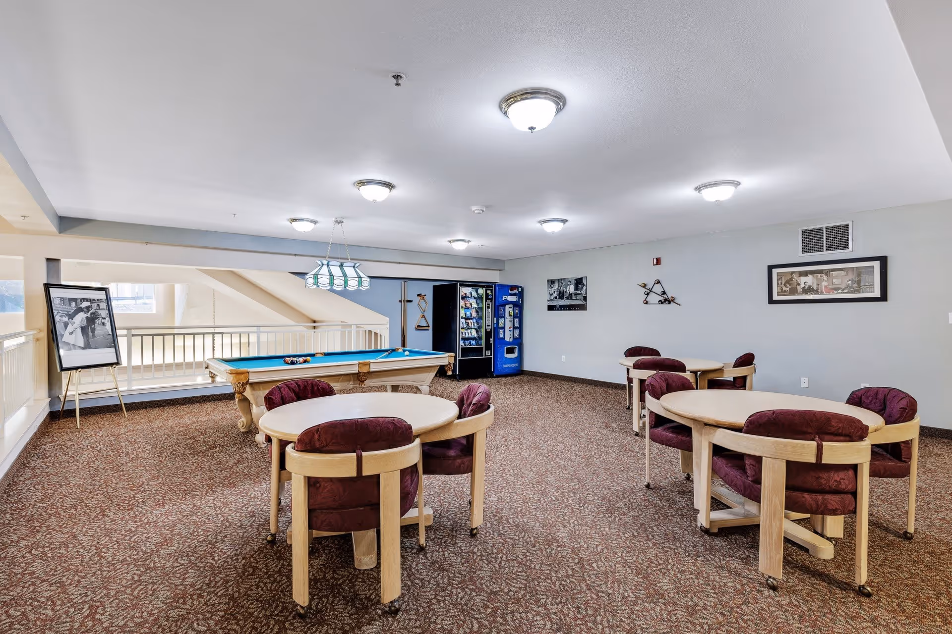 A recreational room with a pool table in the center, surrounded by several round tables with cushioned chairs. There is a vending machine against the far wall, framed pictures hanging on the walls, and a carpeted floor with patterned design. The room is well-lit with ceiling lights and has a railing along one side overlooking a lower level.