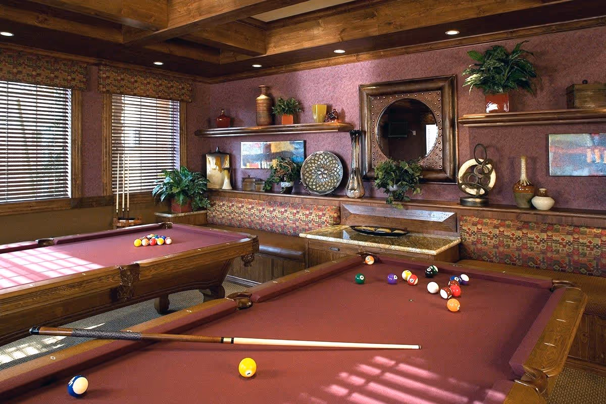 Interior room with two burgundy pool tables, a wooden ceiling with beams, patterned bench seating along the wall, decorative shelves with plants and vases, a round mirror, and windows with wooden blinds letting in sunlight.