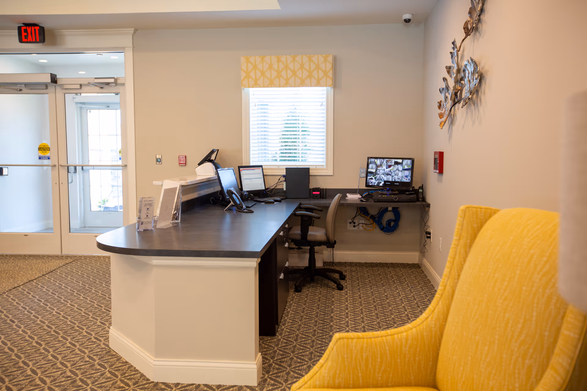 Reception desk area with computers and a security monitor near entry doors and a yellow chair in the foreground.