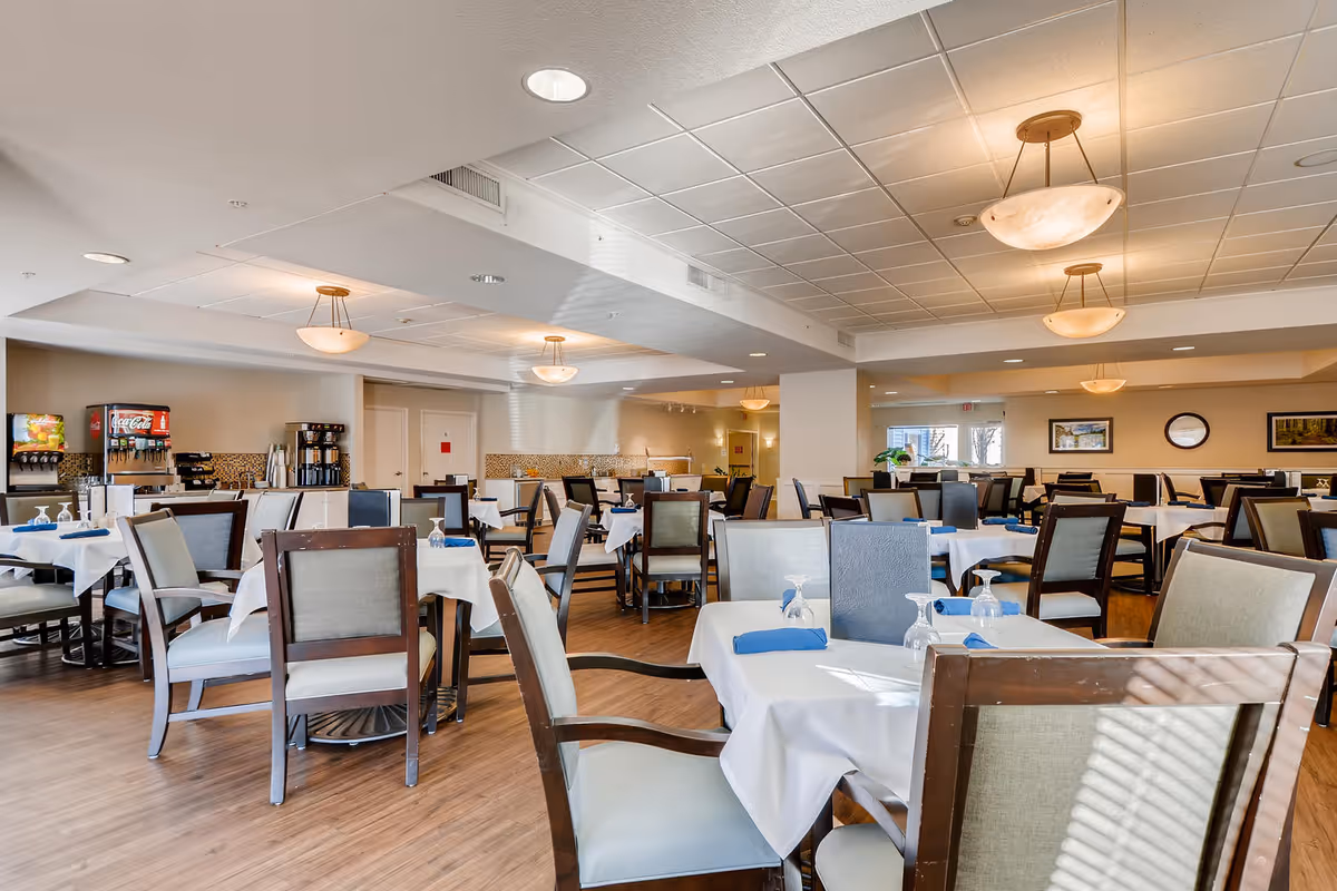 A spacious and well-lit dining room with multiple tables covered in white tablecloths, each set with blue napkins and glassware. The room features wooden chairs with cushioned seats and backs, pendant lights hanging from the ceiling, and a beverage station with coffee and soda machines along one wall. The floor is wooden, and there are framed pictures and a clock on the walls.