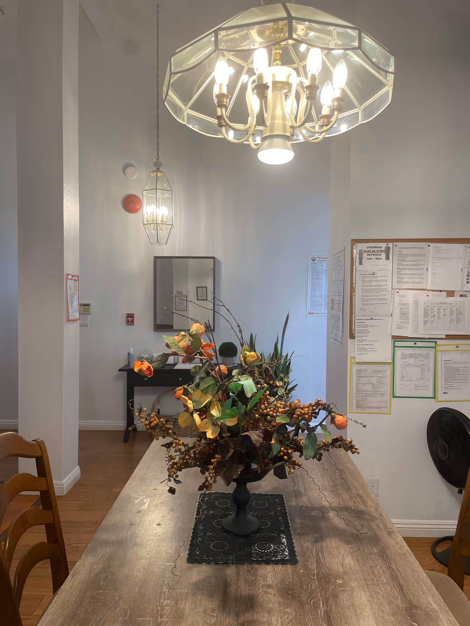 Interior view of a dining area with a wooden table featuring a floral centerpiece. Above the table hangs a chandelier with multiple lit bulbs. In the background, there is a wall with a bulletin board covered in papers, a mirror, and a small table with decorative items. Wooden chairs are partially visible around the table.