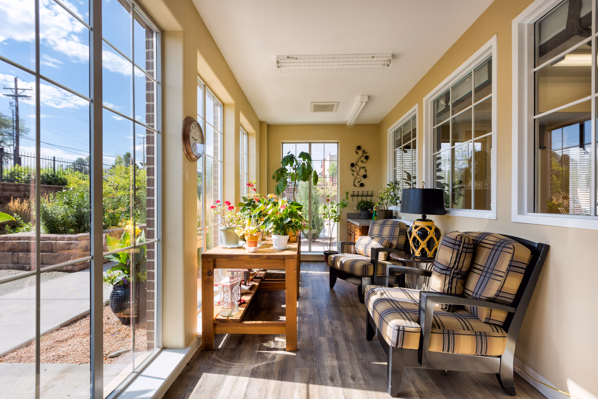 A bright sunroom with large windows letting in natural light, featuring a wooden table with potted plants and two cushioned armchairs with plaid upholstery. The room has light-colored walls, wood flooring, and a black table lamp on a side table between the chairs. Outside the windows, greenery and a clear blue sky are visible.