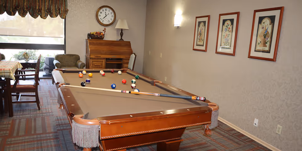 Interior room with a pool table in the center, pool cues resting on the table, and billiard balls arranged on the felt. To the left, there is a dining table with chairs and a window with a valance. In the background, there is a wooden roll-top desk with a lamp and a clock on the wall above it. Three framed floral artworks hang on the right wall.