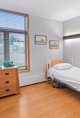 A tidy single-occupancy bedroom with a hospital-style bed, wooden dresser by a window with blinds, and framed pictures on the wall.