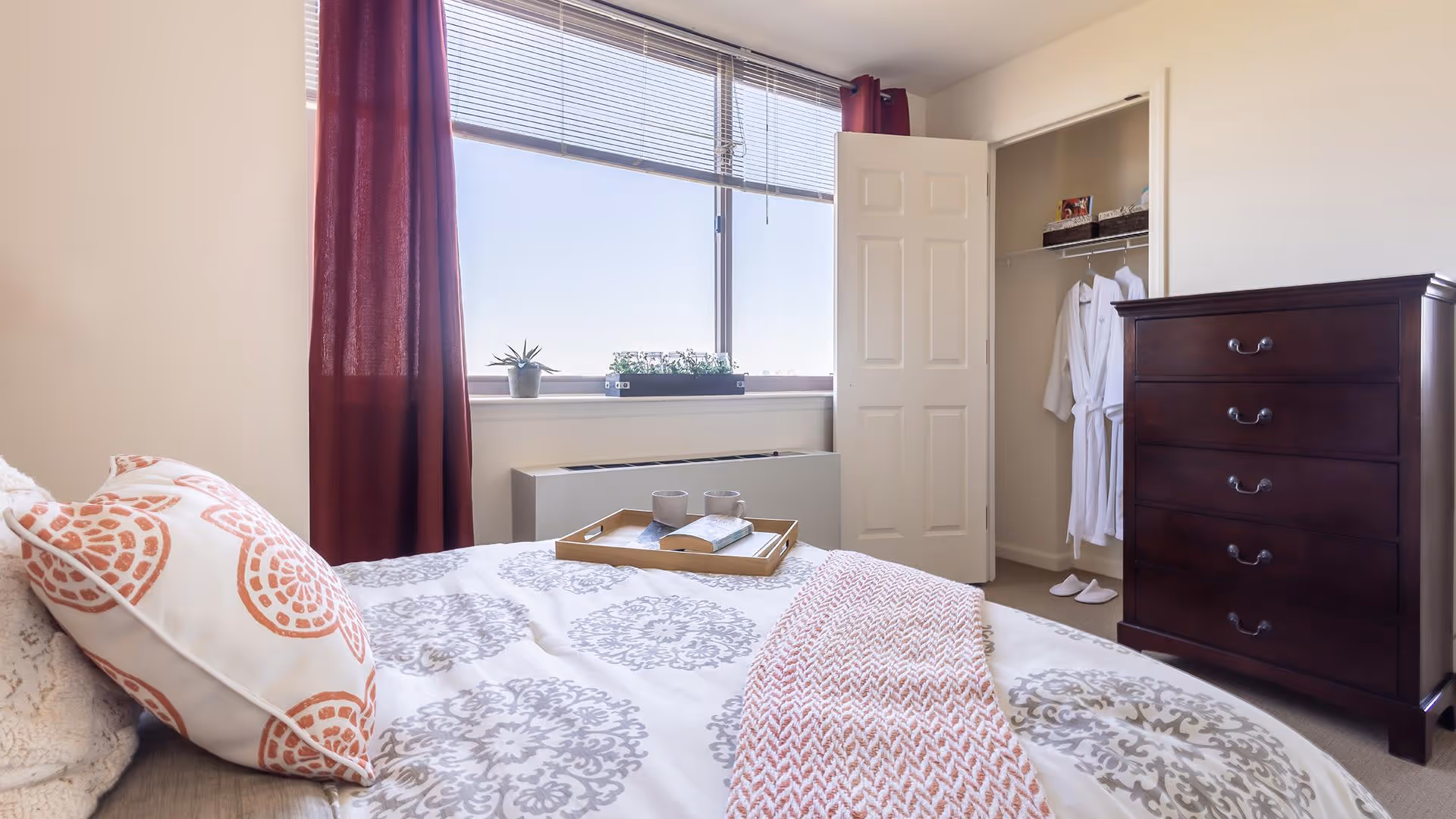 A bright bedroom with a bed featuring patterned pillows and a blanket, a wooden chest of drawers, an open closet with white robes and slippers, and a large window with blinds and burgundy curtains letting in natural light.