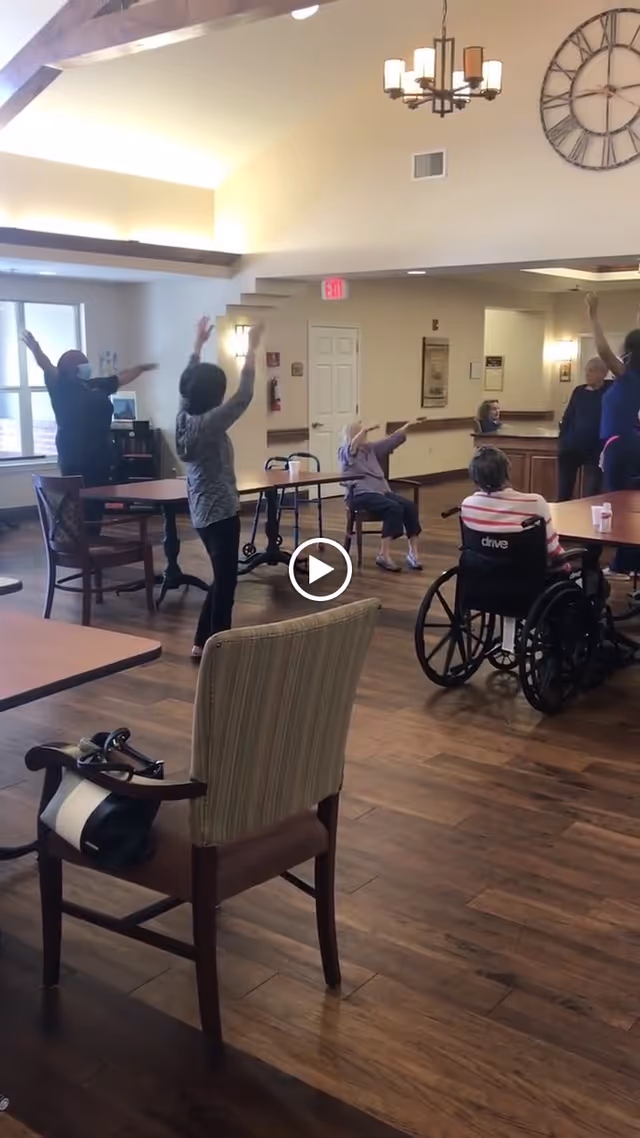 Seniors and staff doing seated and standing group exercises in a spacious activity/common room with wood floors and a vaulted ceiling.