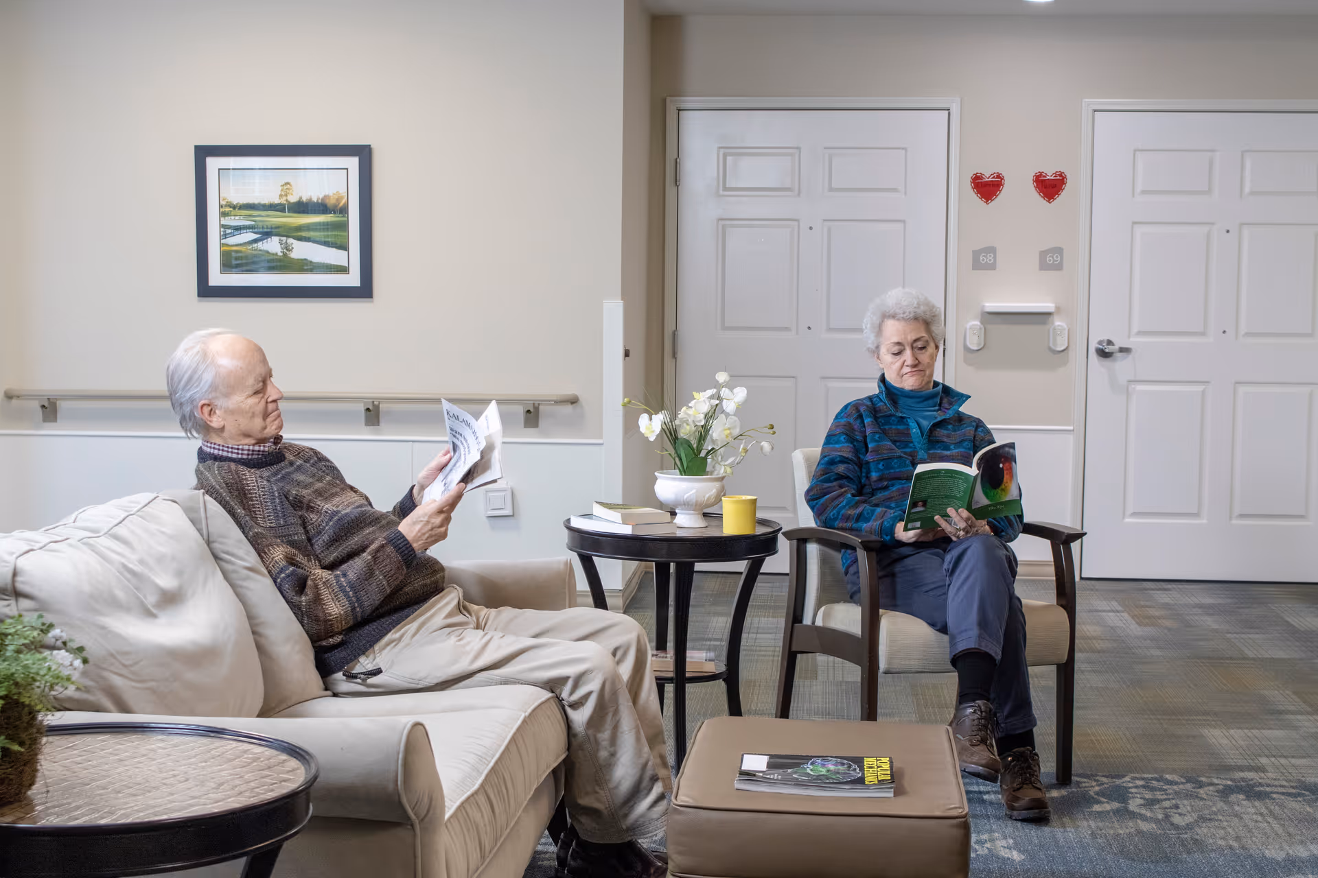 Two elderly individuals sitting in a well-lit common area of a senior living facility. One man is seated on a beige couch reading a newspaper, while a woman sits in a chair reading a book. There are small tables with books, a yellow cup, and a flower arrangement between them. The background shows two white doors with room numbers 68 and 69, and two red heart decorations on the wall.