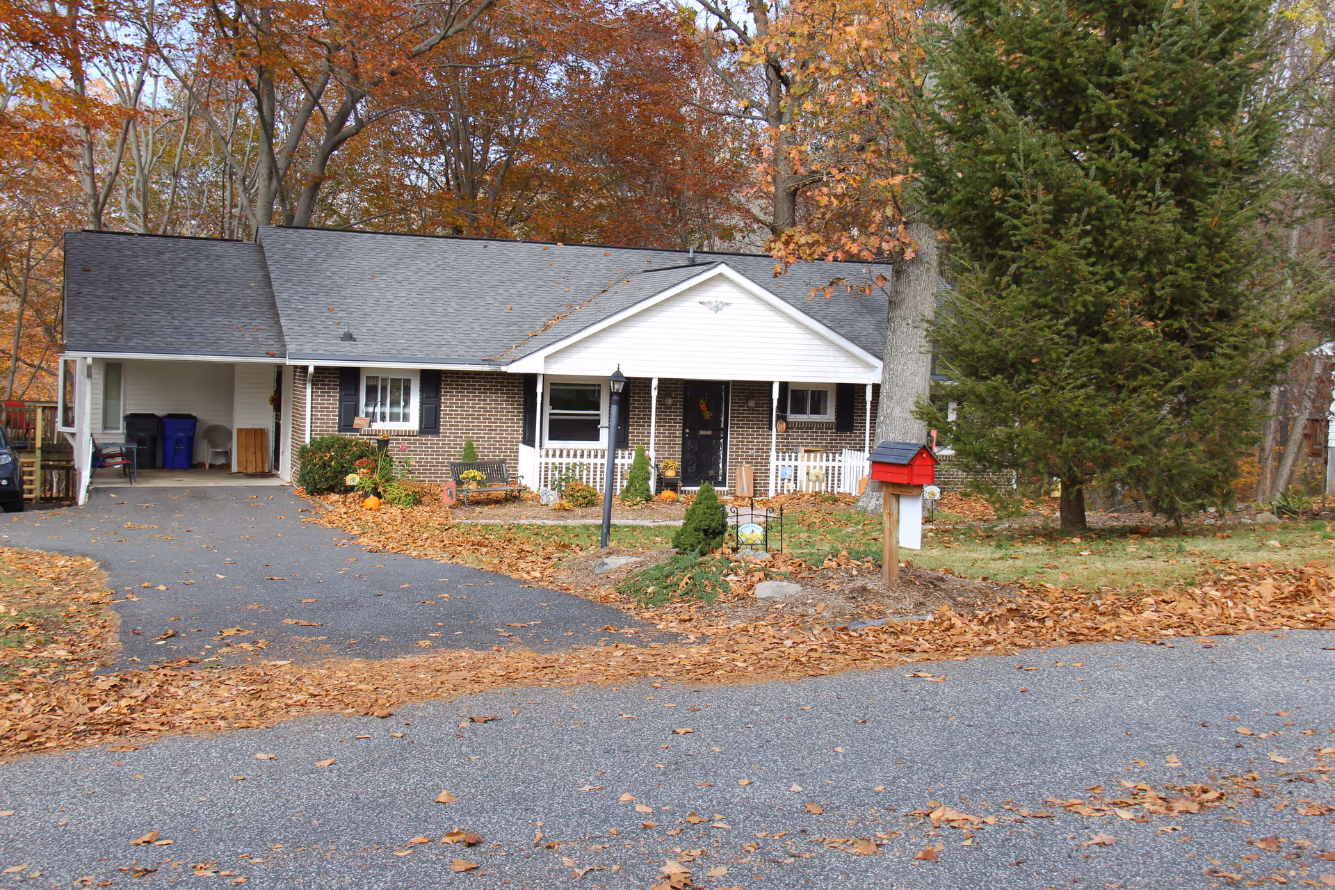 Front exterior view of a single-story brick house with a gray roof, white trim, and a small covered porch. The driveway leads to a carport on the left side. The yard has autumn leaves scattered on the ground, a red mailbox on a wooden post, and various trees with fall foliage in the background.