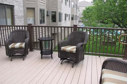 Outdoor patio area with three wicker chairs featuring striped cushions and a small round black table, surrounded by a wooden railing. In the background, there are residential buildings and green trees.