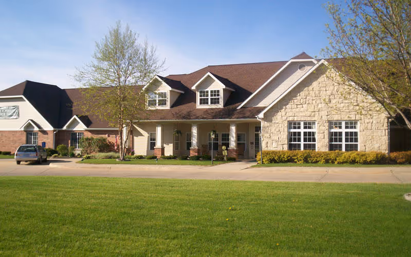 Front exterior of a single-story senior living facility with a porch, parked car, and green lawn.