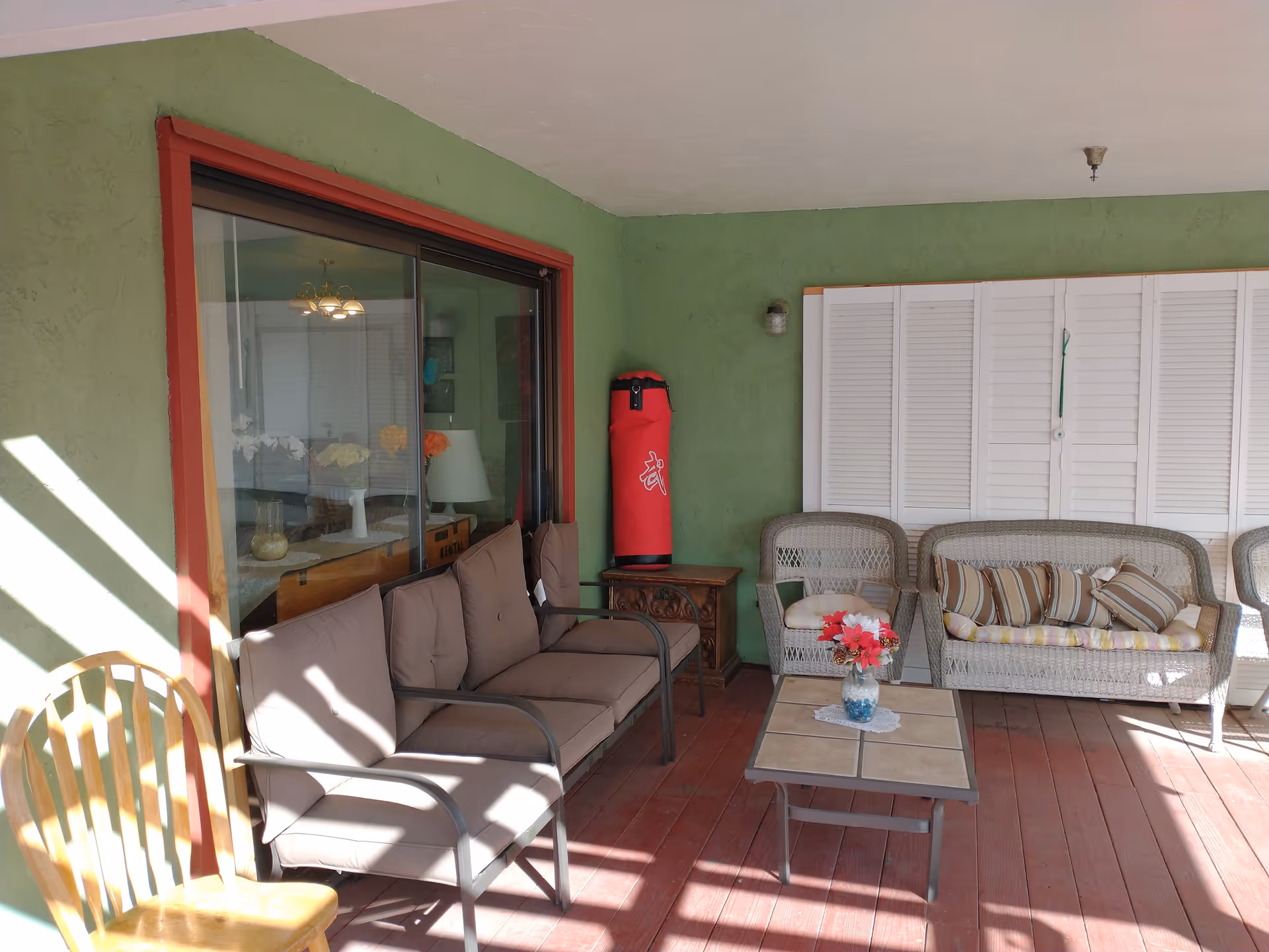 Covered outdoor patio area with green walls and red wooden floor. There is a brown cushioned metal sofa, a wooden chair, a wicker loveseat with striped cushions, and a matching wicker chair. A small table with a vase of flowers is in front of the seating. A red punching bag is placed on a small wooden cabinet against the wall. A large window with a red frame looks into an interior room with a dining table and chandelier.