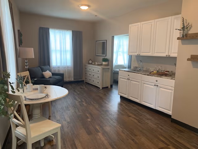 A cozy living space with a small round dining table set for two on the left, a dark gray loveseat near a window with curtains, a white dresser with decorative items, and a kitchenette with white cabinets and a granite countertop on the right. The room has wood flooring and neutral-colored walls.
