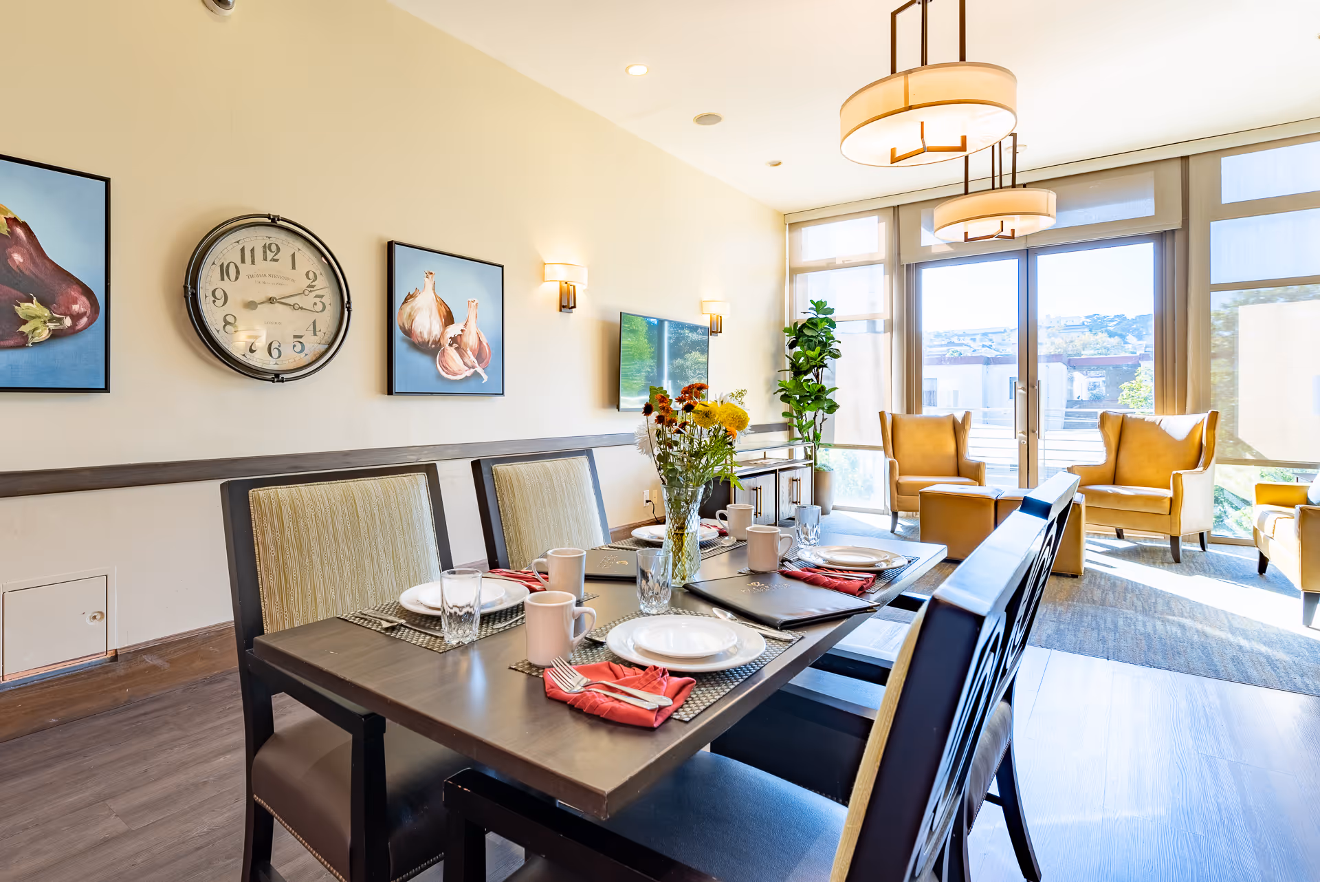 A bright dining area with a dark wooden table set for four, including plates, glasses, mugs, and red napkins. Behind the table are two framed pictures of vegetables and a large wall clock. In the background, there is a living space with two mustard-yellow armchairs, a large window with blinds, and a potted plant. The room is well-lit with natural light and modern ceiling lights.