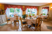 A bright dining room with round tables covered in white tablecloths, each set with plates, cups, and utensils. Wooden chairs with cushions surround the tables. Large windows with red valances let in natural light, and there are plants and decorative items near the windows and walls.