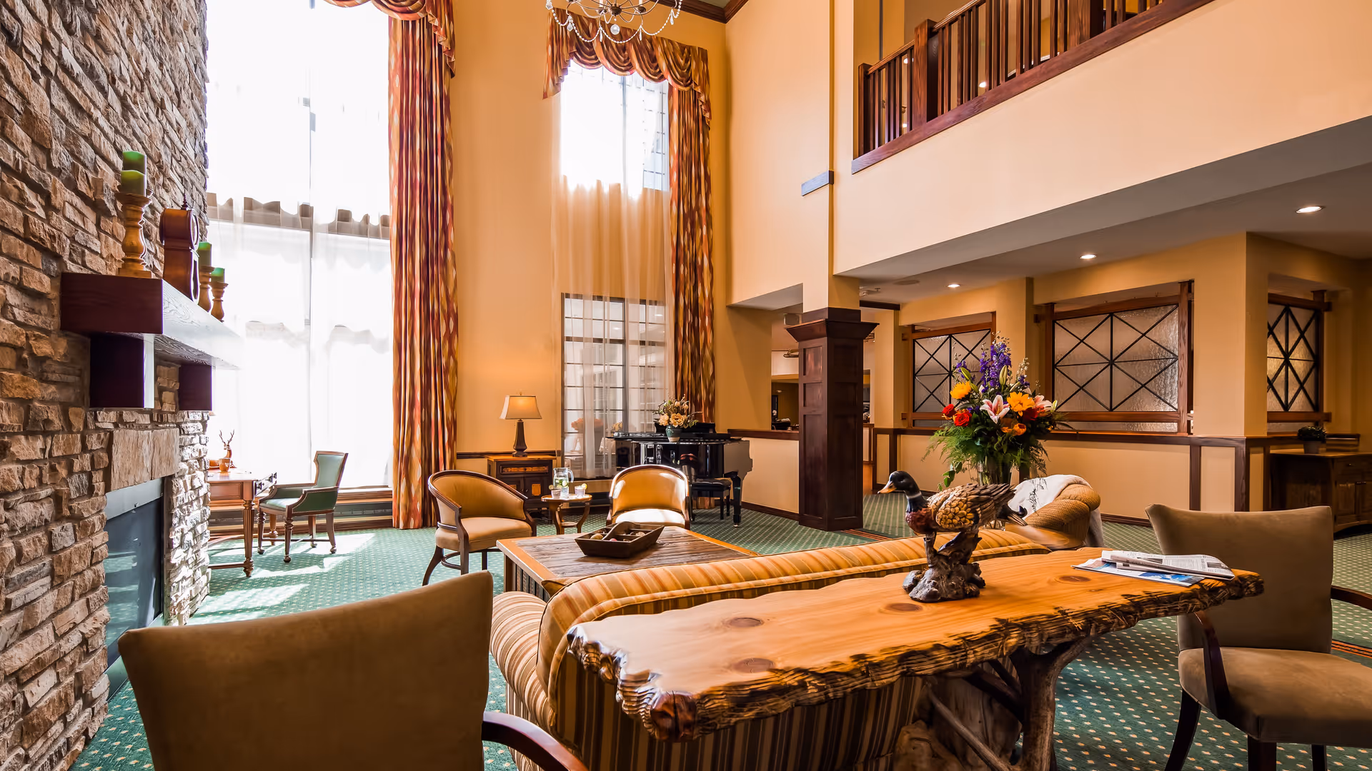 A spacious and well-lit living room area with high ceilings, large windows with long curtains, a stone fireplace, comfortable seating including armchairs and sofas, a wooden coffee table, a decorative wooden console table with a floral arrangement, and a grand piano in the background.