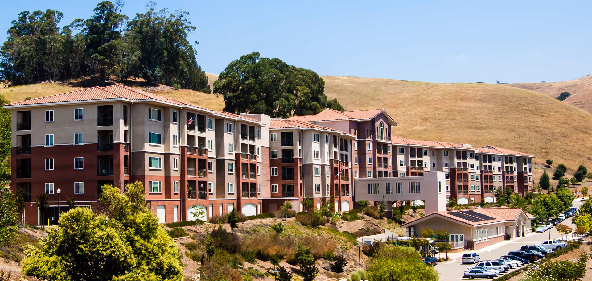 Exterior view of a large multi-story retirement community building with beige and reddish-brown walls and red-tiled roofs, set against dry rolling hills and trees under a clear blue sky. There are parked cars along the road in front of the building.