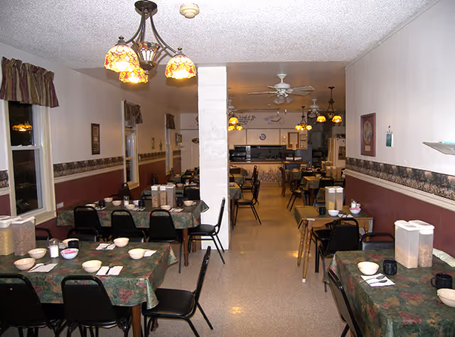 Interior view of a dining room in a senior living facility with multiple tables covered in floral tablecloths, each set with bowls, cups, and napkins. The room has hanging stained glass pendant lights, a ceiling fan, and windows with valances. The walls are painted white with a maroon lower half and decorative border. The room extends into a kitchen area in the background.