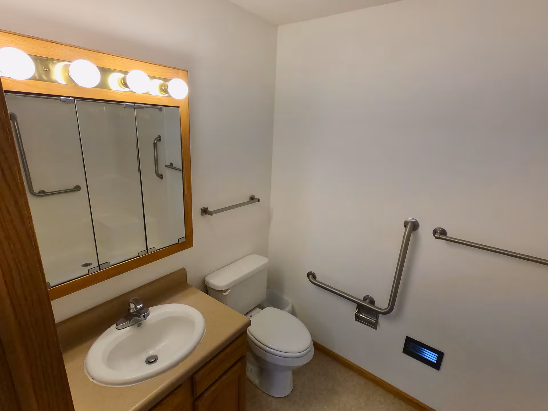 A small bathroom with a white sink set in a beige countertop with wooden cabinetry below. Above the sink is a large mirror with three panels and four round light bulbs mounted on a wooden frame. To the right of the sink is a white toilet. The walls are plain white with several metal grab bars installed, indicating accessibility features. There is a small vent near the floor on the right wall.