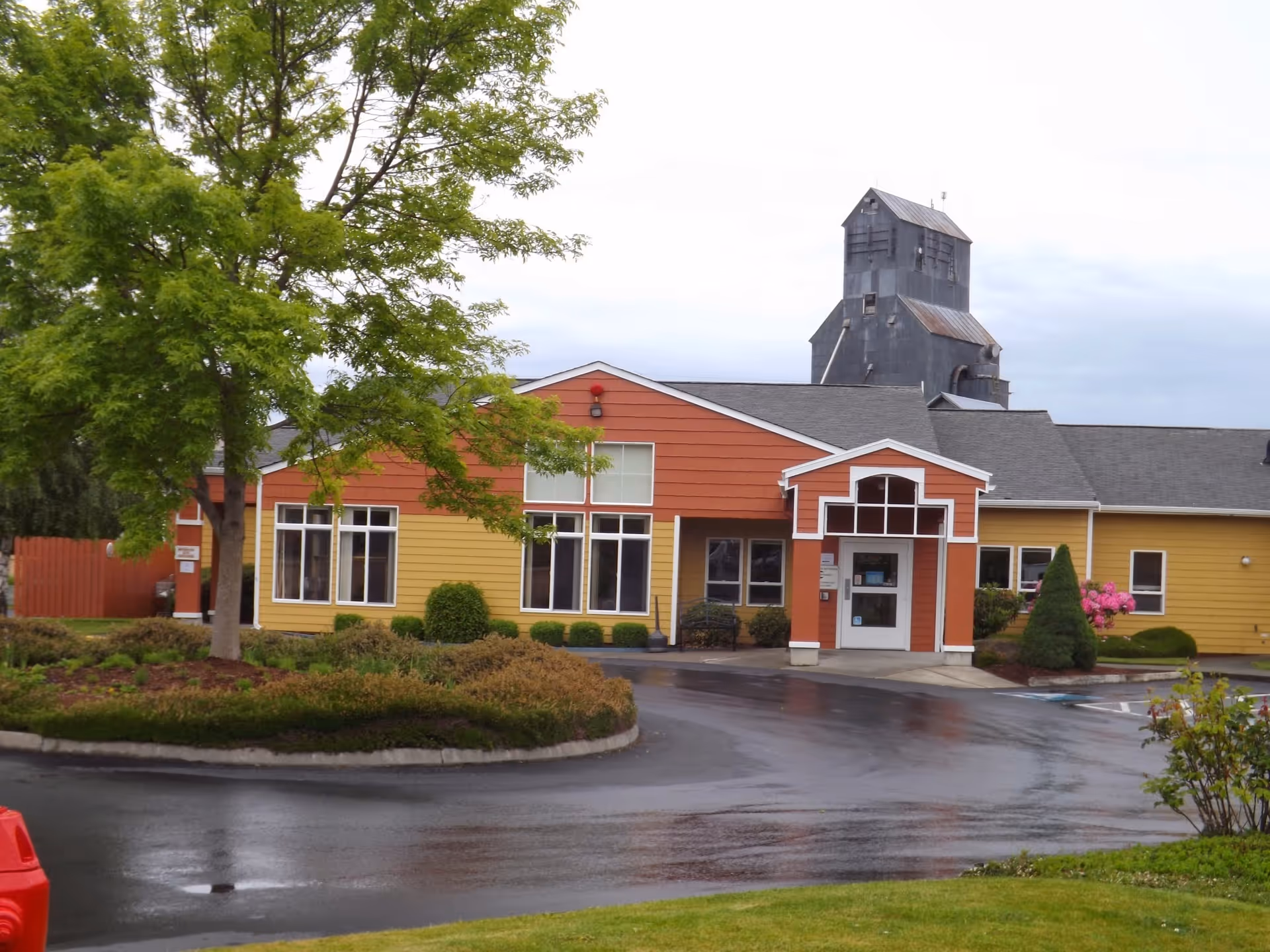 Exterior view of a single-story building with yellow and orange siding, large windows, and a covered entrance. There is a tree and landscaped bushes in front, with a paved driveway and parking area. A tall, dark industrial structure is visible in the background under a cloudy sky.