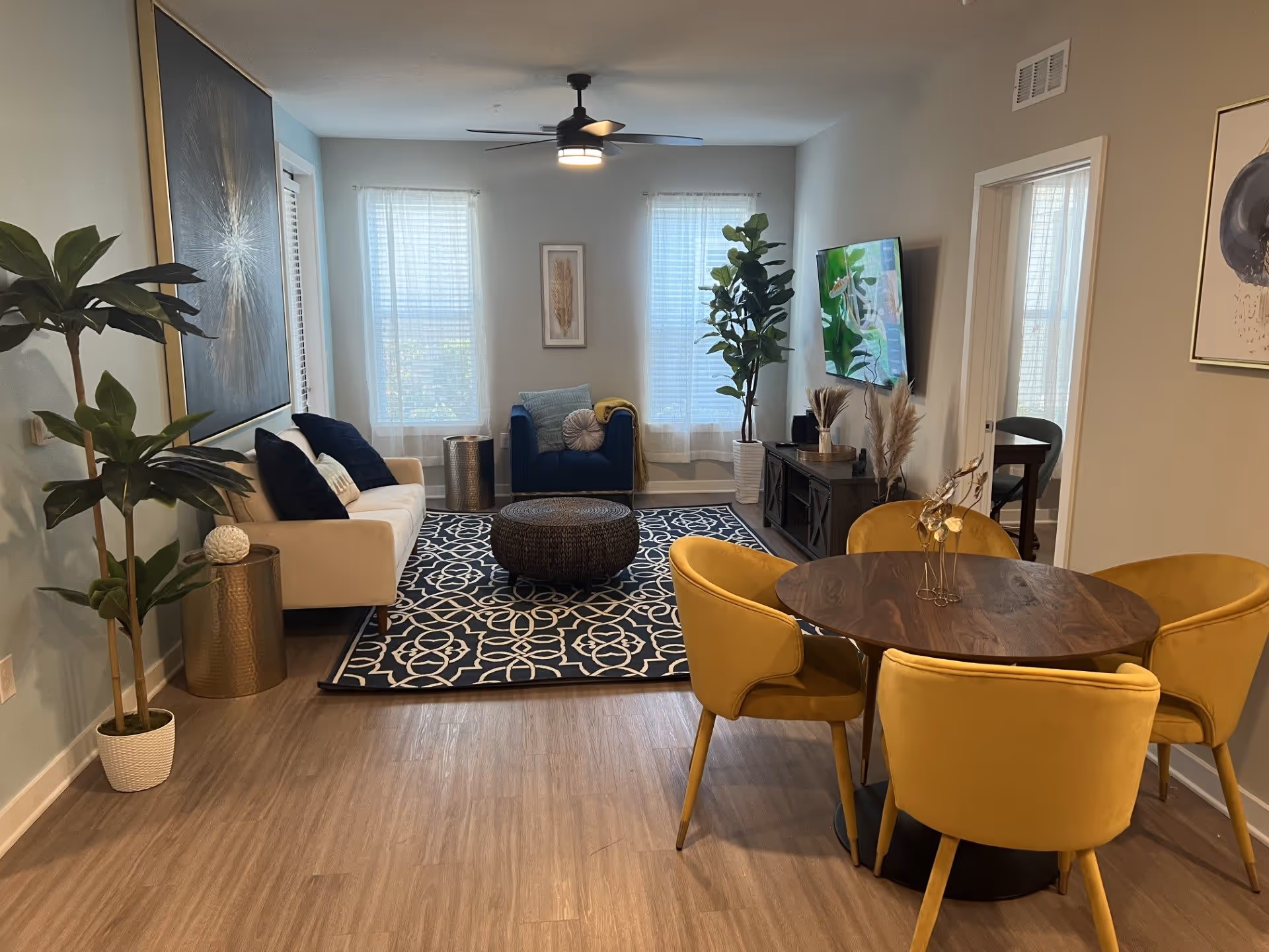 A modern living room and dining area with light wood flooring. The living room features a beige sofa with dark blue cushions, a dark blue armchair, a round woven ottoman on a patterned rug, and a flat-screen TV mounted on the wall. There are two large windows with sheer white curtains, a ceiling fan with light, and several decorative plants. The dining area has a round wooden table surrounded by four mustard yellow upholstered chairs. The walls are painted light gray with framed artwork.