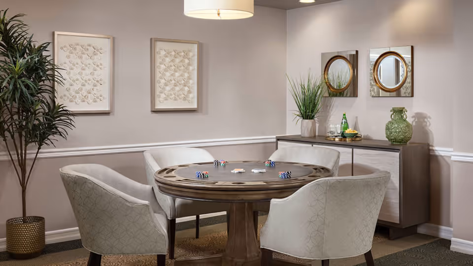 A cozy interior room with a round wooden table set for a card game with poker chips and cards. Four upholstered chairs surround the table. The room features neutral-colored walls with two framed artworks, a tall potted plant in a gold pot, and a sideboard with decorative items including a green vase, a plant, and a tray with glasses and a bottle. Two round mirrors hang above the sideboard.