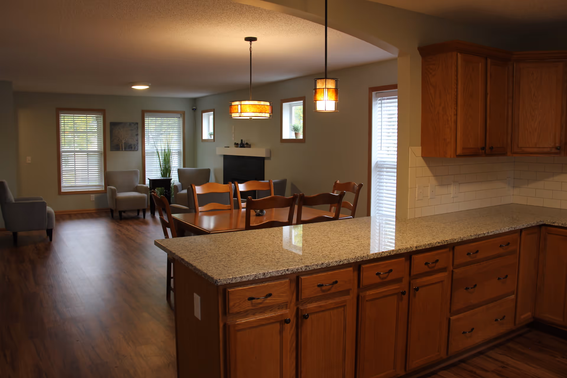 Open-concept kitchen with a granite countertop island and wooden cabinets opening onto a dining table and living area with chairs and windows.