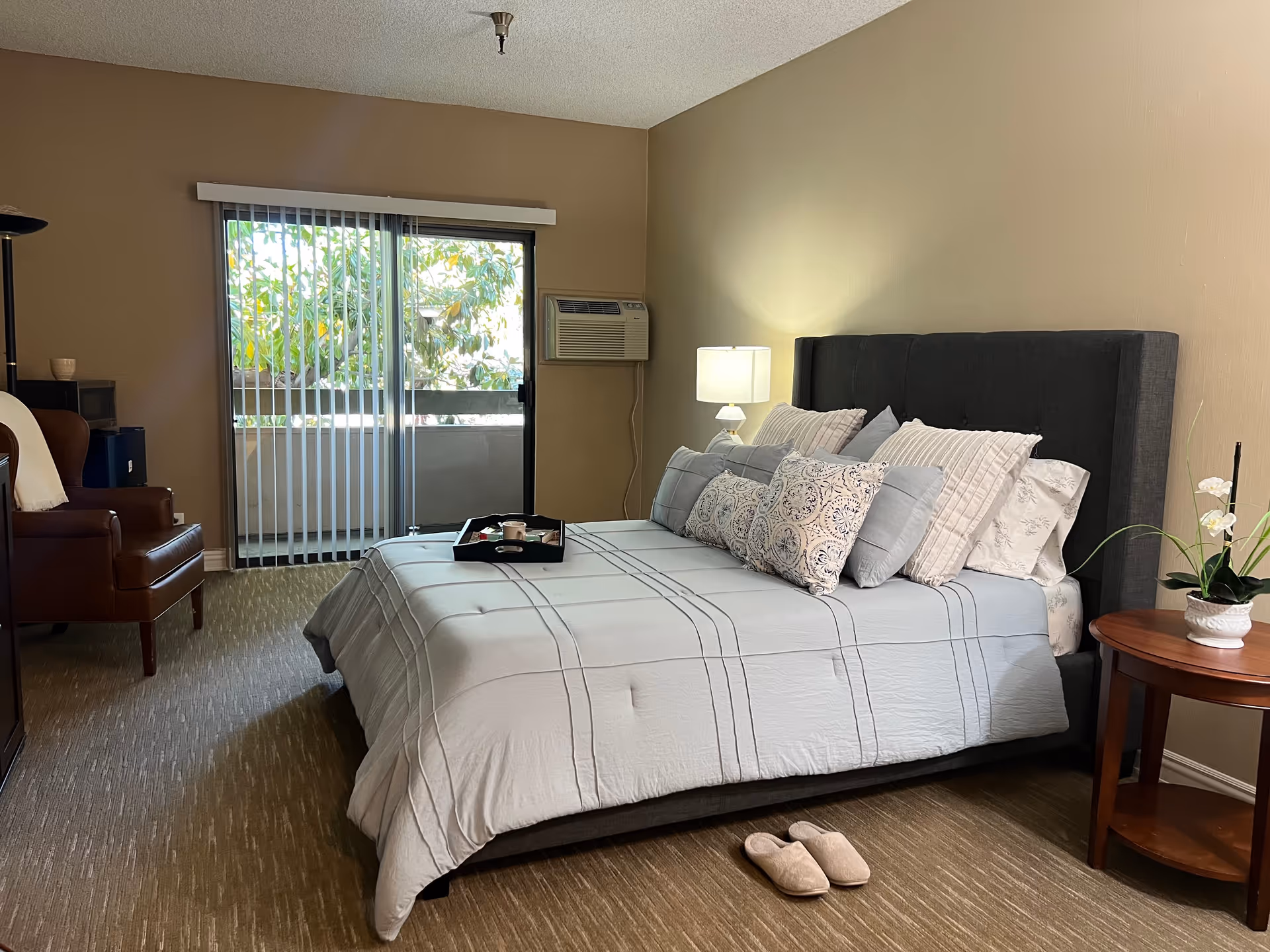 A cozy bedroom with a large bed covered in a light gray comforter and multiple pillows. There is a wooden side table with a white lamp and a potted plant. A pair of slippers is placed on the carpeted floor near the bed. A sliding glass door with vertical blinds leads to a small balcony with greenery visible outside. An air conditioning unit is mounted on the wall next to the door. A brown leather chair and a floor lamp are positioned in the corner of the room.