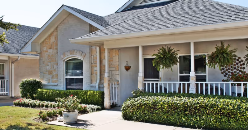 Exterior view of a single-story senior living facility building with a covered porch, white railing, hanging plants, and well-maintained landscaping including bushes and flowers.