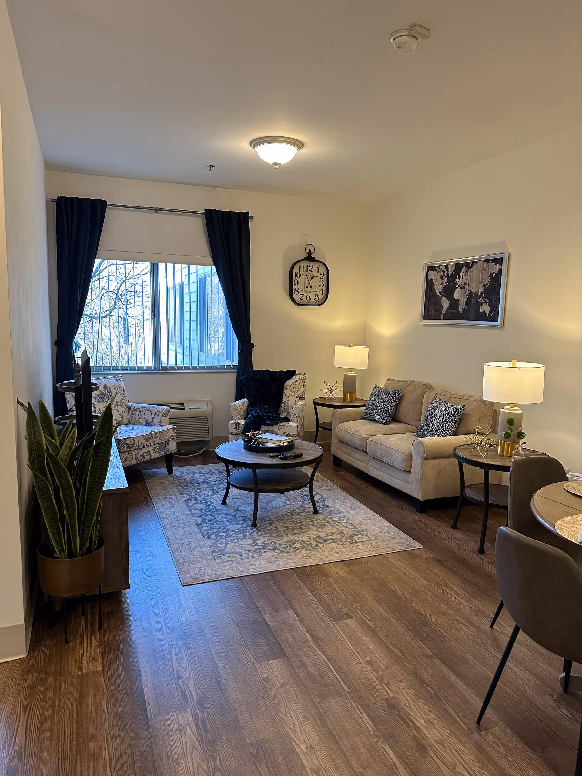 A cozy living room with wooden flooring, a beige sofa with two patterned cushions, two floral armchairs near a window with dark blue curtains, a round coffee table on a patterned rug, two side tables with lamps, a wall clock, a framed world map, and a potted plant near a TV stand.
