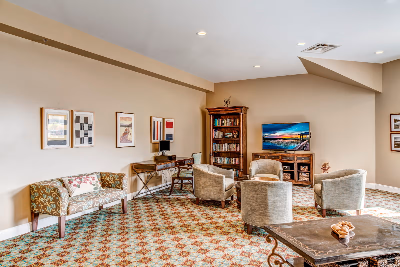 A cozy living room area with patterned carpet, a floral upholstered loveseat, four beige armchairs arranged around a small round wooden table, a wooden bookshelf filled with books, a flat-screen TV on a wooden cabinet displaying a sunset over a pier, a wooden desk with a chair and computer, and framed artwork on the beige walls.